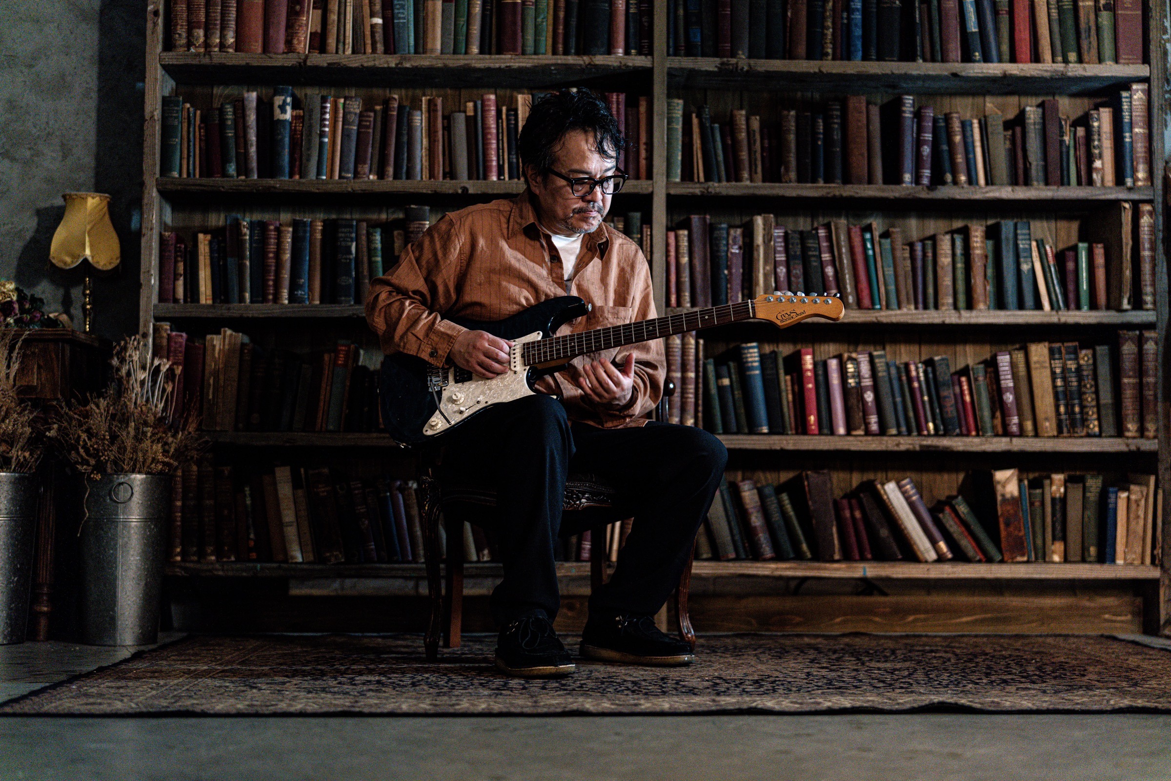 Kenji Suzuki playing electric guitar, seated in front of bookshelves